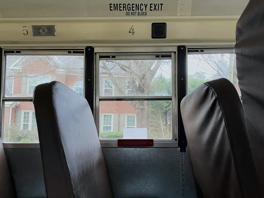 Empty high-back seats inside a school bus beneath an EMERGENCY EXIT sign, with houses visible through the windows
