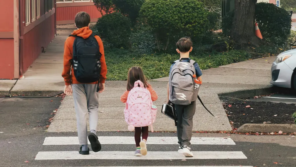 Three children wearing backpacks walking across a crosswalk, seen from behind